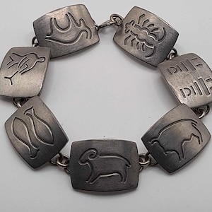 以下が含まれることがあります： 長方形のリンクに干支のシンボルが刻まれたシルバーカラーのブレスレット。牡羊座、蠍座、その他の占星術のサインのシンボルが含まれています。リンクは小さなチェーンリンクで接続されています。