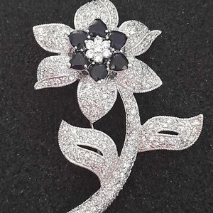 May include: A silver flower brooch with a black centre and clear rhinestones. The flower has five petals and a stem with leaves.