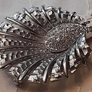May include: A silver-toned brooch with a textured, fan-like design. The centre of the brooch features a cluster of rhinestones.