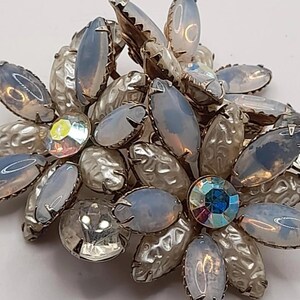 May include: A vintage brooch with a floral design. The brooch features opalescent, oval-shaped stones and textured silver-tone metal petals. A large, iridescent crystal sits at the center, surrounded by smaller crystals.