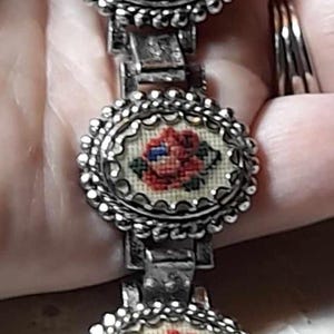 Pode incluir: Um bracelete de prata com quatro pingentes redondos. Cada pingente apresenta um fundo branco com uma rosa vermelha bordada no centro. Os pingentes são conectados por uma corrente de prata.