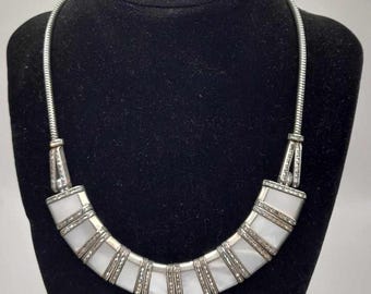 Puede incluir: Un conjunto de collar y pendientes plateados. El collar presenta un colgante curvo, blanco y rectangular, con detalles plateados y una cadena plateada. Los pendientes son rectangulares con un centro blanco y un borde plateado.