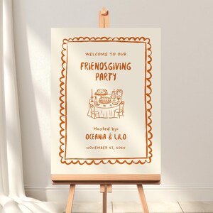 May include: A cream-colored Friendsgiving party sign on a wooden easel. The sign has a decorative brown border and text that reads "Welcome to our Friendsgiving Party" with an illustration of a table setting. The sign is hosted by Oceania & Lilo, November 17, 2015.