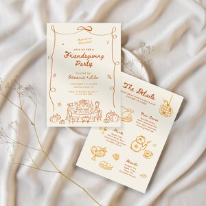 May include: Two cream-colored Friendsgiving party invitations with orange illustrations and text. One card reads "Join us for a Friendsgiving Party" and the other lists the menu. The cards are on a white fabric background.
