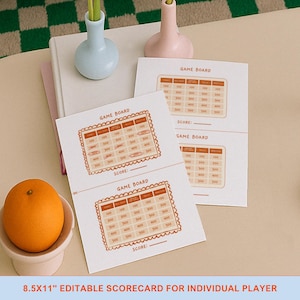 Op de afbeelding: Witte spelscorecards met een oranje rand en een rasterindeling. De kaarten hebben de tekst "GAME BOARD" en "SCORE:" erop gedrukt. Een sinaasappel ligt in een kleine witte kom. De tekst "8.5x11" EDITABLE SCORECARD FOR INDIVIDUAL PLAYER" staat onderaan.