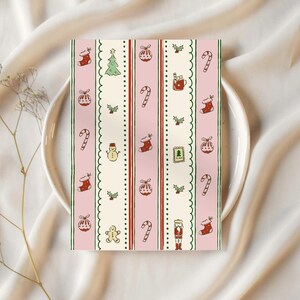 May include: A rectangular card with a pink background and vertical stripes featuring Christmas-themed illustrations. The design includes candy canes, stockings, ornaments, a Christmas tree, and a snowman. The card is placed on a white plate.
