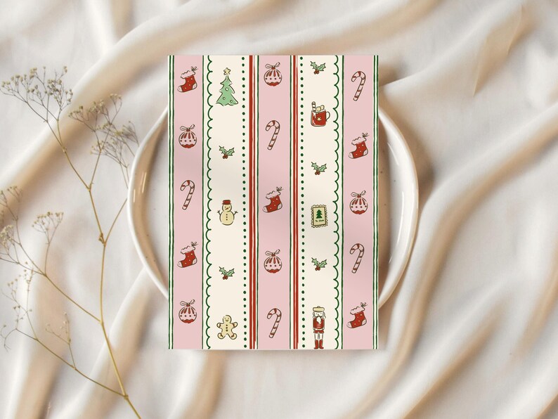 May include: A rectangular card with a pink background and vertical stripes featuring Christmas-themed illustrations. The design includes candy canes, stockings, ornaments, a Christmas tree, and a snowman. The card is placed on a white plate.