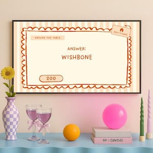Op de afbeelding: Een ingelijst spelbeeld met het antwoord "WISHBONE". Een vaas met een zonnebloem, twee wijnglazen met roze strikken, een roze ballon, een oranje bal, boeken en kaarsen staan op een blauwe tafel.