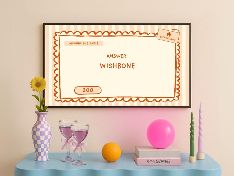 Op de afbeelding: Een ingelijst spelbeeld met het antwoord "WISHBONE". Een vaas met een zonnebloem, twee wijnglazen met roze strikken, een roze ballon, een oranje bal, boeken en kaarsen staan op een blauwe tafel.