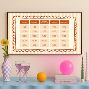 Op de afbeelding: Een ingelijst trivia-spelbord met categorie&euml;n en puntwaarden. Een vaas met een zonnebloem, twee wijnglazen met roze strikken, een roze ballon, een oranje bol, twee boeken en twee kaarsen staan op een blauwe tafel.