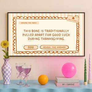 Op de afbeelding: Een ingelijst Thanksgiving-spel met de tekst "This bone is traditionally pulled apart for good luck during Thanksgiving." De sc&egrave;ne omvat een vaas met een zonnebloem, wijnglazen en een roze ballon.