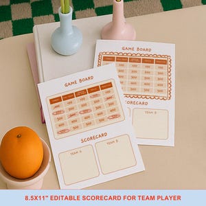 Op de afbeelding: Twee scorekaarten voor een spelbord met een raster van getallen en de tekst "GAME BOARD". De scorekaarten liggen op een lichtbeige oppervlak, met een sinaasappel in een kleine witte kom op de voorgrond.