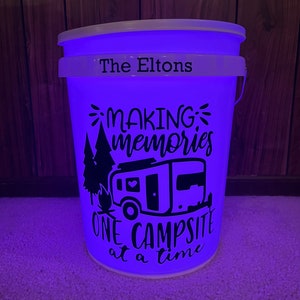 May include: A white bucket with a lid and handle, illuminated with a blue glow. The text "The Eltons" is above the phrase "MAKING memories ONE CAMPSITE at a time", along with a camper and trees.
