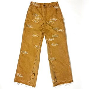 May include: Brown work pants with multiple white embroidered name patches. The pants have a classic workwear design with a straight leg, multiple pockets, and a button and zipper closure. The names are in a cursive font.