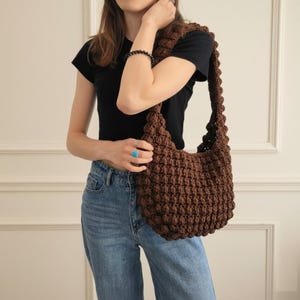 May include: A brown, hand-crocheted shoulder bag with a textured, bubble-like pattern. The bag has a wide strap and a rounded shape. The bag is made of a soft, brown yarn and is worn over the shoulder.