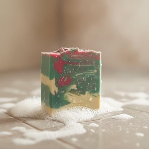 May include: A rectangular bar of soap featuring a marbled swirl pattern in shades of deep green, cream, and magenta. The top surface has a textured finish with small dark particles, and the base is surrounded by a thick layer of white soap lather.