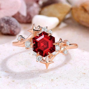 May include: Rose gold ring featuring a large, hexagonal, deep red gemstone at the center, surrounded by smaller white gemstones and star-shaped accents. The ring is set against a neutral background, highlighting its intricate design.