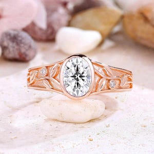 May include: Rose gold ring with an oval-cut, clear gemstone center. The band features a leaf design with small, round accent stones. The ring is displayed on a white stone, with a blurred background of other stones and flowers.