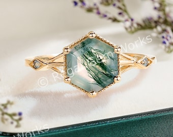 Puede incluir: Un anillo de oro con una piedra preciosa de ágata verde hexagonal y dos pequeños diamantes en la banda. El anillo está en una caja de terciopelo verde.