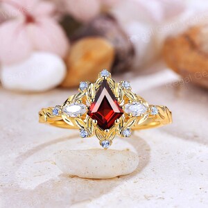 May include: A gold ring with a central, kite-shaped, deep red gemstone, surrounded by white and light blue accent stones. The ring has an ornate, floral design with intricate detailing. The ring is displayed on a light-coloured surface.