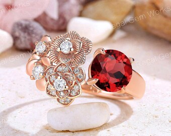 May include: A rose gold ring with a large red gemstone surrounded by smaller white gemstones. The ring features a floral design with three delicate flowers.