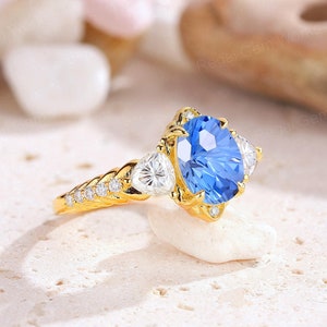 May include: A gold engagement ring with a large oval blue gemstone surrounded by smaller clear gemstones. The ring has a braided band.