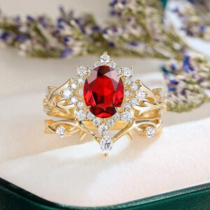Könnte beinhalten: Ein goldfarbenes Ring-Set mit einem großen, oval geschliffenen roten Edelstein, umgeben von einem Halo aus kleineren weißen Edelsteinen. Das Band hat ein filigranes, verziertes Design mit zusätzlichen kleinen weißen Edelsteinen. Das Set enthält ein passendes Band.