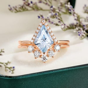 May include: A rose gold engagement ring set with a large, light blue, square-shaped center stone surrounded by smaller diamonds. The ring is set in a delicate, prong setting and has a halo of smaller diamonds around the center stone. The ring is in a green velvet ring box.