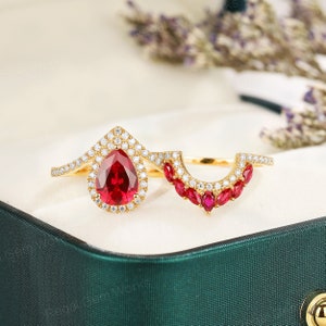 Op de afbeelding: Een verlovingsring met een peervormige robijn en een halo van kleinere diamanten. De ring is gezet in geelgoud en heeft een bijpassende trouwring met een rij kleinere robijnen.