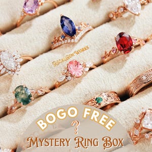 May include: A collection of rose gold rings with various gemstone settings, including sapphires, rubies, and emeralds. The rings are displayed on a cream-colored surface. The image includes the text "BOGO FREE Mystery Ring Box".