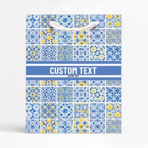 Op de afbeelding: Vierkante cadeauzak met een blauw en geel tegelpatroon. De tas heeft een wit lint handvat en een blauwe band met de tekst "CUSTOM TEXT". Ideaal voor cadeaus, met een decoratief tegelpatroon.