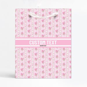 Puede incluir: Una bolsa de regalo rosa con un estampado floral de pequeñas rosas rosas y rayas verticales blancas. La bolsa tiene una banda rosa con las palabras "CUSTOM TEXT" impresas en blanco. Asas de cuerda blanca.