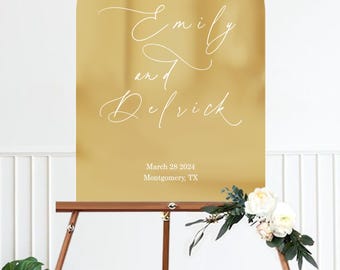 May include: A gold-colored wedding welcome sign with an arched top, displayed on a wooden easel. The sign reads "Welcome to our Wedding" and features the names "Emily and Derrick" in elegant script, along with the wedding date and location.