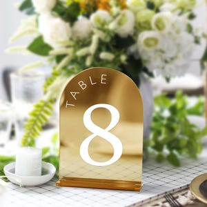 May include: A gold-colored table number with the word "TABLE" and the number "8" in white. The table number is arched at the top and sits on a small wooden base. The background is blurred, showing flowers and a candle.