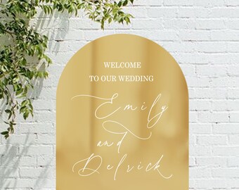 May include: A gold arched wedding sign with white script that reads "Welcome to our wedding" and the names "Emily and Delrick". The date and location are also displayed. The sign is on a wooden easel, in front of a white brick wall.