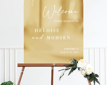 May include: A gold-colored wedding welcome sign with an arched top, displayed on a wooden easel. The sign reads "Welcome" and lists the names "Heloise and Mohsen" along with the wedding date and location.