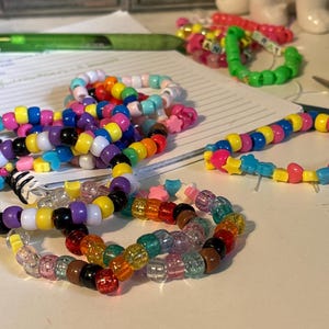 May include: A close-up of colorful beaded bracelets with star and heart charms. The bracelets are made with small plastic beads in various colors, including yellow, blue, pink, purple, and white. The bracelets are arranged on a white surface.
