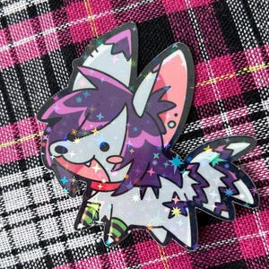 May include: A colorful enamel pin featuring a cartoon-style creature with purple, white, and pink accents. The pin has a holographic finish and is set against a pink and black plaid background.