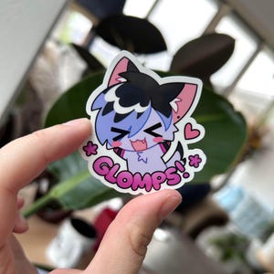 Op de afbeelding: Een sticker met een cartoonfiguur met blauwe en paarse vacht, zwart haar en een roze hart. Het figuur lacht en er staat "*GLOMPS!*" onder.