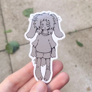 Może przedstawiać: Szara naklejka z postacią z kreskówki królika z długimi uszami, ubranego w sweter i szorty. Królik ma lekko zmęczony wyraz twarzy. Naklejka jest trzymana na rozmytym tle zewnętrznym.