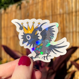 May include: A holographic sticker of a black bird wearing a gold crown. The bird has a rainbow iridescent pattern on its body and wings.