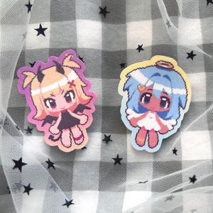 Peut inclure: Deux autocollants de personnages de dessins animés, l'un avec un fond rose et un personnage de diable avec des cornes et une queue, et l'autre avec un fond bleu et un personnage d'ange avec des ailes et une auréole.
