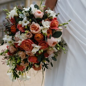 May include: A bridal bouquet with peach, orange, and white flowers. The bouquet is held by a person wearing a white dress.