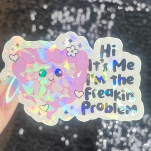 May include: Holographic sticker with a pink brain-shaped design, featuring a smiling face and hearts. The sticker has the text "Hi It's Me I'm the Freakin Problem" in a playful font. The sticker has a shiny, iridescent finish.