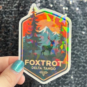 May include: A holographic sticker with a mountain and forest scene, featuring a deer. The sticker has the text "FOXTROT DELTA TANGO" and a butterfly graphic. The sticker is in a hexagonal shape with a white border.