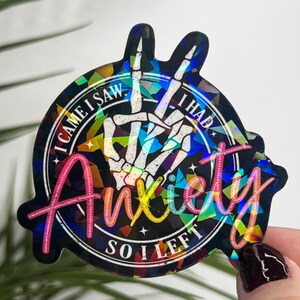 K&ouml;nnte beinhalten: Ein holografischer Aufkleber mit einer Skeletthand, die das Peace-Zeichen macht. Der Aufkleber zeigt den Text "I came I saw I had anxiety so I left" in einer Regenbogen-Schrift.