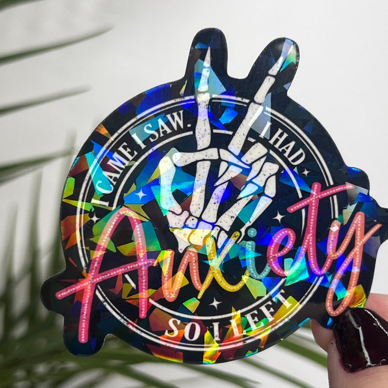K&ouml;nnte beinhalten: Ein holografischer Aufkleber mit einer Skeletthand, die ein Peace-Zeichen macht. Der Aufkleber hat einen schwarzen Hintergrund mit einem bunten, glitzernden Design. Der Text auf dem Aufkleber lautet "I came I saw I had anxiety so I left."