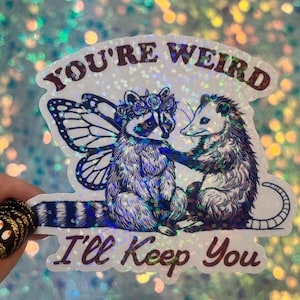 May include: A holographic sticker featuring a raccoon with butterfly wings and a possum. The raccoon wears a flower crown. Text reads "You're Weird, I'll Keep You" in a burgundy color. The sticker has a glittery, iridescent finish.