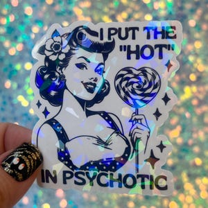 May include: Holographic sticker featuring a retro-style illustration of a woman with dark hair and a flower, holding a heart-shaped lollipop. Text reads "I PUT THE 'HOT' IN PSYCHOTIC" in bold, uppercase letters. The sticker has a shiny, iridescent finish.