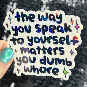 May include: Holographic sticker with the text "the way you speak to yourself matters you dumb whore" in a handwritten style. The sticker has a white background with colorful star accents.